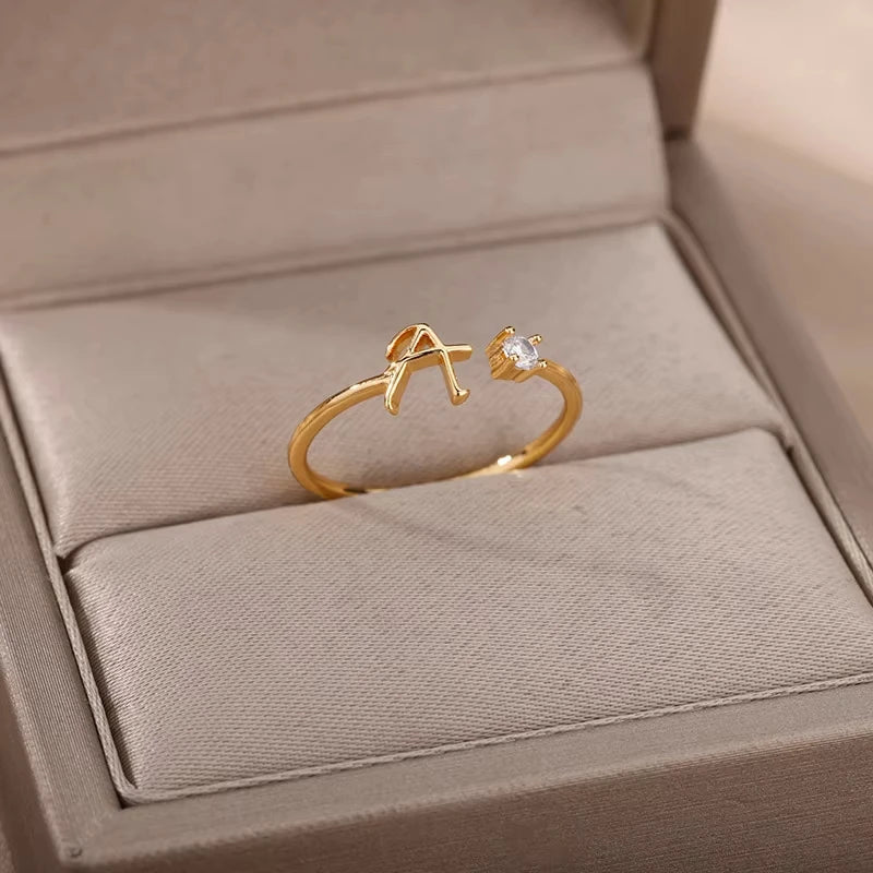 Infinity elegant gold ring featuring a delicate open band design with a small sparkling crystal on one end and a stylish, artistic "A" shape on the other, presented in a soft beige jewelry box.