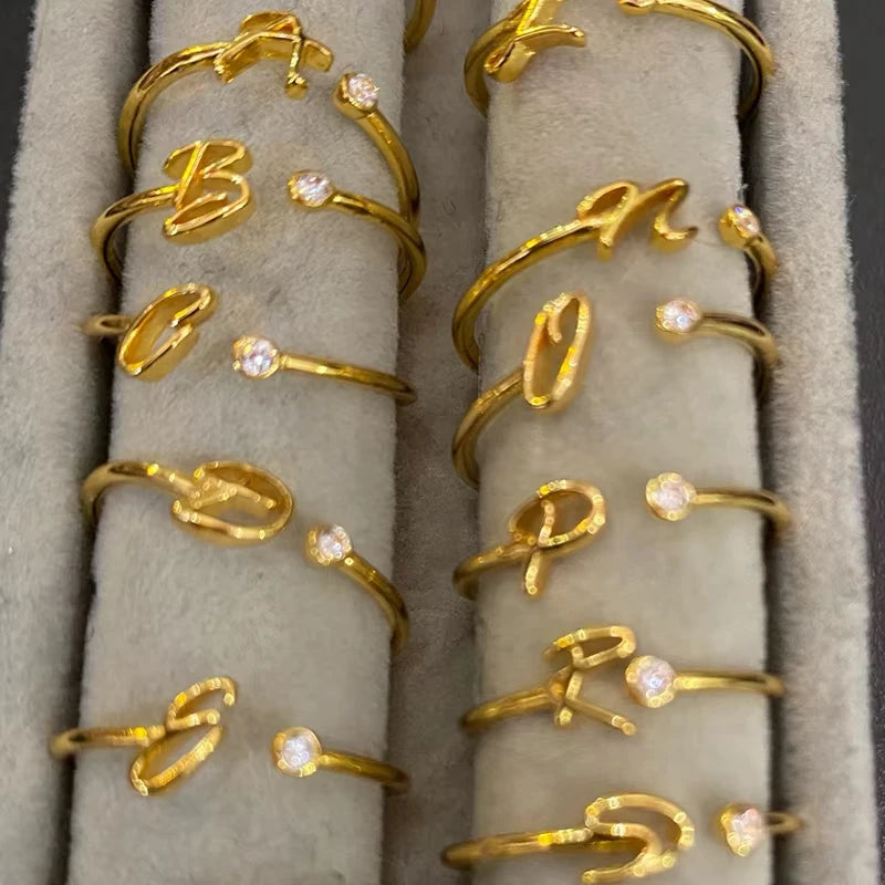 A collection of elegant gold rings from the Infinity series, each featuring a delicate script letter design paired with a small sparkling crystal, displayed on a soft beige jewelry holder.