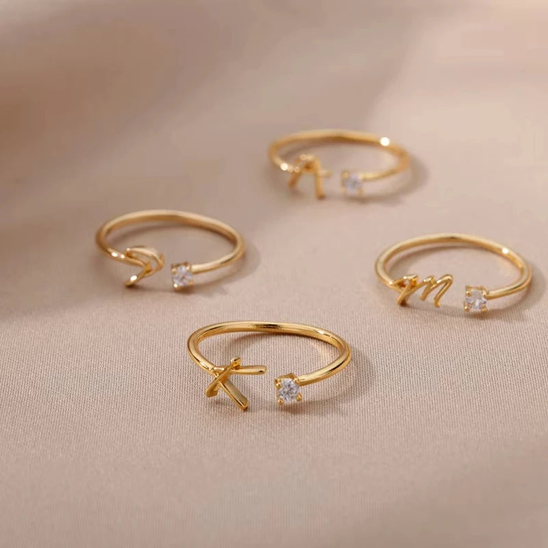 A set of delicate gold rings from the Infinity collection, each featuring a minimalist band with a small sparkling crystal and an elegant cursive letter detail, embodying timeless luxury and modern sophistication.